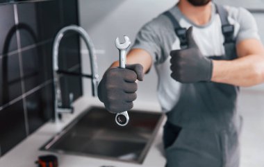 Close up view of young professional plumber in grey uniform holding wrench in hand on the kitchen.
