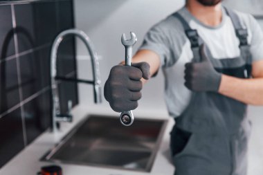 Close up view of young professional plumber in grey uniform holding wrench in hand on the kitchen.