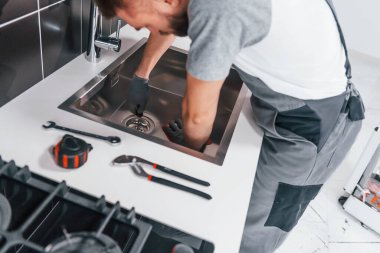 Young professional plumber in grey uniform working on the kitchen.