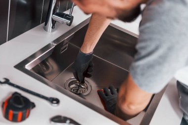 Young professional plumber in grey uniform working on the kitchen.