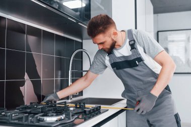 Young professional plumber in grey uniform measuring table by using meter on the kitchen.