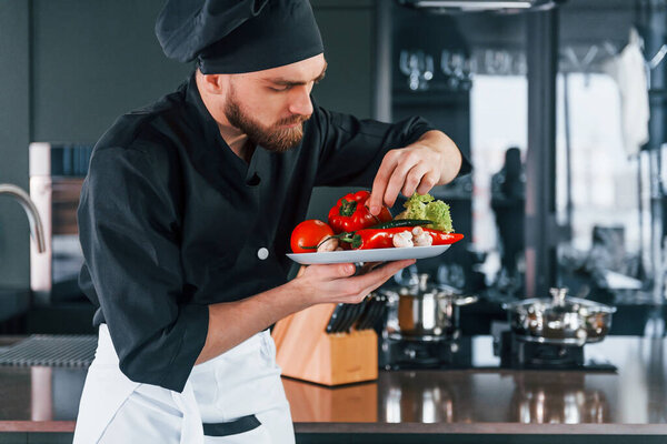 Professional young chef cook in uniform holds plate with vegetables on the kitchen.
