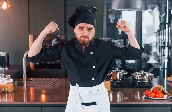 Professional young chef cook in uniform standing near table and posing for a camera on the kitchen.