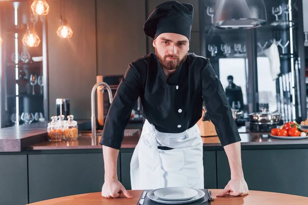 Profesyonel aşçı üniforması içinde masanın yanında duruyor ve mutfakta kameraya poz veriyor..
