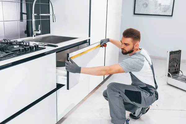 Gri üniformalı genç profesyonel tesisatçı. Mutfakta taksimetre kullanarak ölçü alıyor..