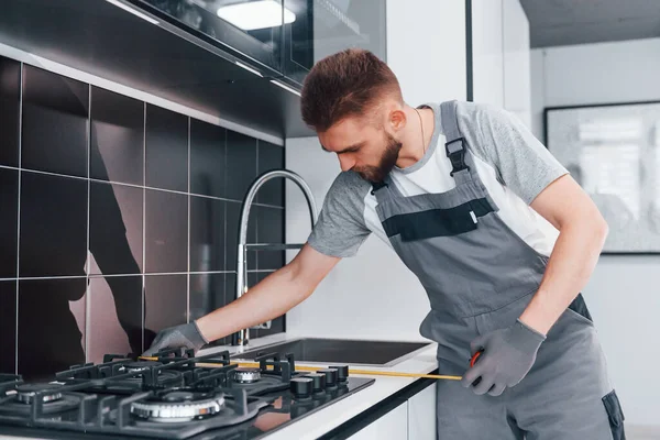 Gri üniformalı genç profesyonel tesisatçı. Mutfakta taksimetre kullanarak ölçü alıyor..