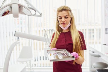 Üniformalı genç kadın dişçi stomatoloji ofisinde elinde aletlerle dikiliyor..