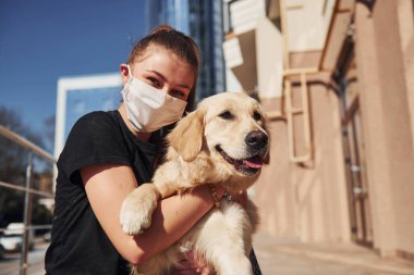 Sitting together. Young positive woman in protective mask have a walk outdoors near business building at quarantine time. Conception of coronavirus.