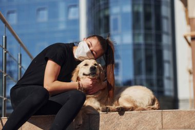 Birbirinize sarılın. Korunaklı maskeli genç bir kadın köpeğiyle birlikte karantina koronavirüsünde oturuyor..