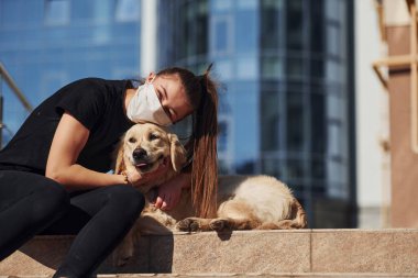 Birbirinize sarılın. Korunaklı maskeli genç bir kadın köpeğiyle birlikte karantina koronavirüsünde oturuyor..