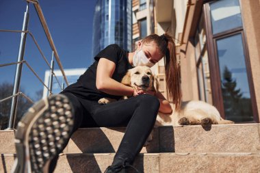 Birbirinize sarılın. Korunaklı maskeli genç bir kadın köpeğiyle birlikte karantina koronavirüsünde oturuyor..