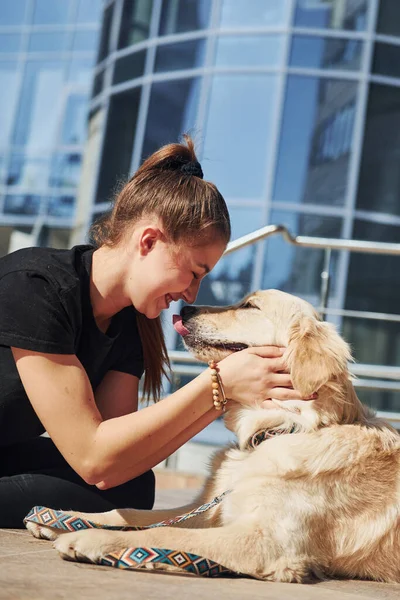 Young positive woman have fun with her dog when have a walk outdoors near business building.