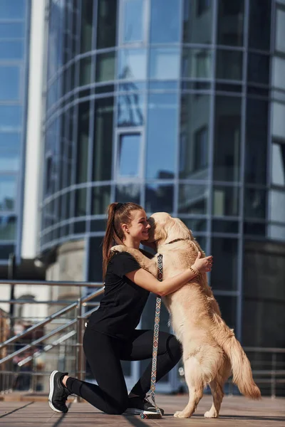 Young positive woman have fun and doing tricks with her dog when have a walk outdoors near business building.