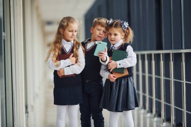 School kids in uniform together with phone in corridor. Conception of education.