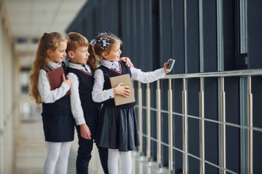 School kids in uniform together with phone and making selfie in corridor. Conception of education.
