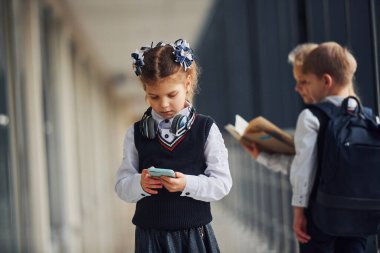 Little girl with phone standing if front of school kids in uniform that together in corridor. Conception of education.