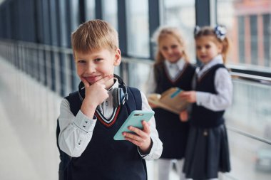 Little boywith phone and headphones standing if front of school kids in uniform that together in corridor. Conception of education.