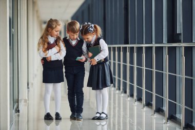 School kids in uniform together with phone in corridor. Conception of education.