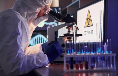 Masadaki bilgili monitörler. Beyaz koruyucu üniformalı bilim adamları laboratuarda koronavirüs ve kan tüpleriyle çalışıyorlar..
