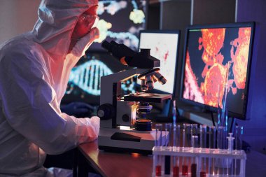 Monitors with information on the table. Scientist in white protective uniform works with coronavirus and blood tubes in laboratory.
