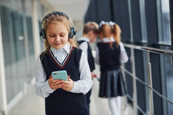 廊下で一緒に制服を着た学校の子供たちの前に立っている携帯電話やヘッドフォンを持つ少女 教育の概念 — ストック写真