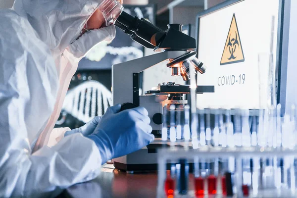 Masadaki bilgili monitörler. Beyaz koruyucu üniformalı bilim adamları laboratuarda koronavirüs ve kan tüpleriyle çalışıyorlar..