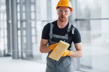 Gri üniformalı bir adamın portresi. Gündüz vakti modern bir ofiste elinde ekipmanla ayakta duruyor..