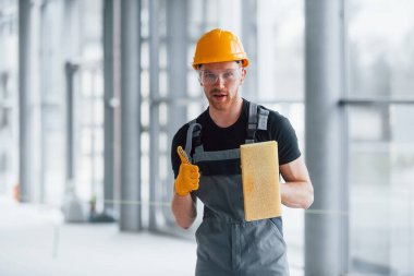 Gri üniformalı bir adamın portresi. Gündüz vakti modern bir ofiste elinde ekipmanla ayakta duruyor..