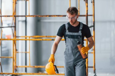 Man in grey uniform near construction takes break indoors in modern big office at daytime.