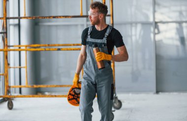 Man in grey uniform near construction takes break indoors in modern big office at daytime.