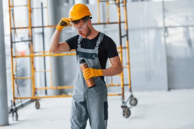 Man in grey uniform near construction takes break indoors in modern big office at daytime.