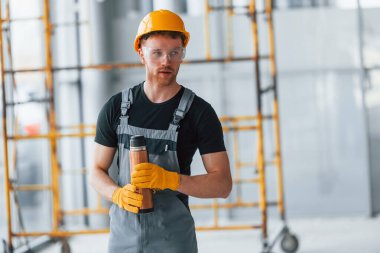 Man in grey uniform near construction takes break indoors in modern big office at daytime.