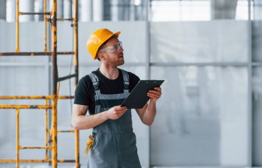 Young engineer in grey uniform with notepad works indoors in modern big office at daytime.