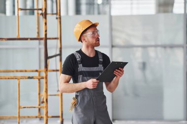 Young engineer in grey uniform with notepad works indoors in modern big office at daytime.