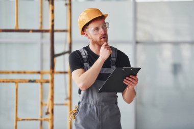 Young engineer in grey uniform with notepad works indoors in modern big office at daytime.