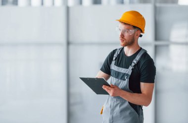 Young engineer in grey uniform with notepad works indoors in modern big office at daytime.