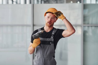 Gri üniformalı ve elinde matkapla modern büyük ofiste gündüz vakti ayakta duran bir adam..