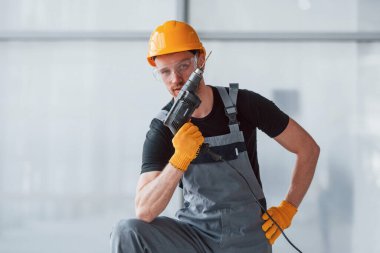 Gri üniformalı ve elinde matkapla modern büyük ofiste gündüz vakti ayakta duran bir adam..