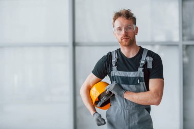 Gri üniformalı ve elinde turuncu kasketli bir adam portresi. Gündüz vakti büyük bir ofiste kapalı duruyor..