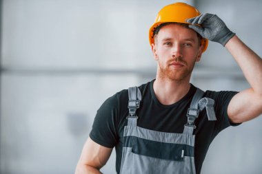 Portrait of man in grey uniform and orange hard hat that standing indoors in modern big office at daytime.