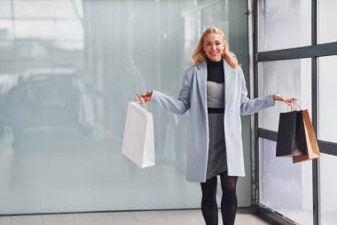 Beautiful blonde in elegant clothes with packages standing indoors against grey background.