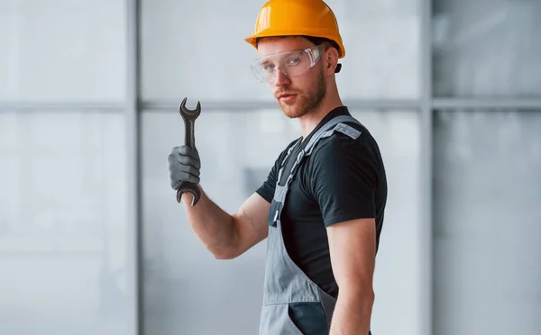 Man in grey uniform with wrench in hand standing indoors in modern big office at daytime.
