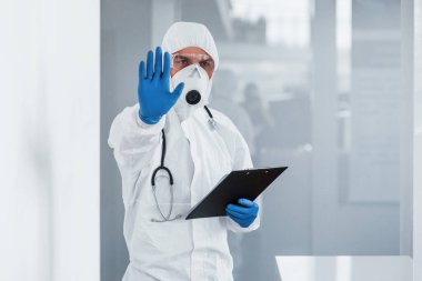 Shows stop gesture by hand. Male doctor scientist in lab coat, defensive eyewear and mask holding notepad in hands.