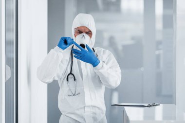Laboratuvar önlüklü, koruyucu gözlüklü ve maskeli bir erkek doktor mavi eldivenlerini çıkarıyor..