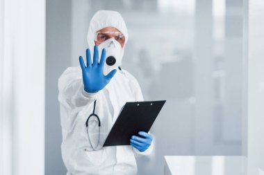 Shows stop gesture by hand. Male doctor scientist in lab coat, defensive eyewear and mask holding notepad in hands.