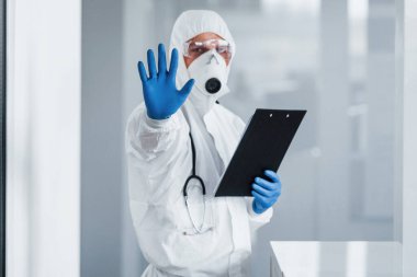 Shows stop gesture by hand. Male doctor scientist in lab coat, defensive eyewear and mask holding notepad in hands.