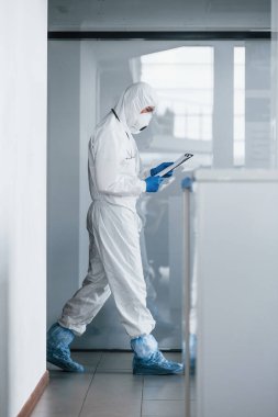Laboratuvar önlüklü, koruyucu gözlüklü ve maskeli bir erkek doktor elinde not defteri tutuyor..