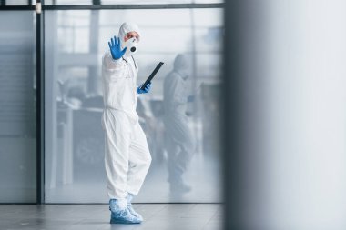 Shows stop gesture by hand. Male doctor scientist in lab coat, defensive eyewear and mask holding notepad in hands.
