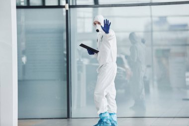 El hareketlerini durdurur. Laboratuvar önlüklü, koruyucu gözlüklü ve maskeli bir erkek doktor elinde not defteri tutuyor..