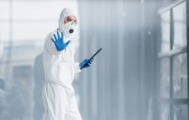 El hareketlerini durdurur. Laboratuvar önlüklü, koruyucu gözlüklü ve maskeli bir erkek doktor elinde not defteri tutuyor..